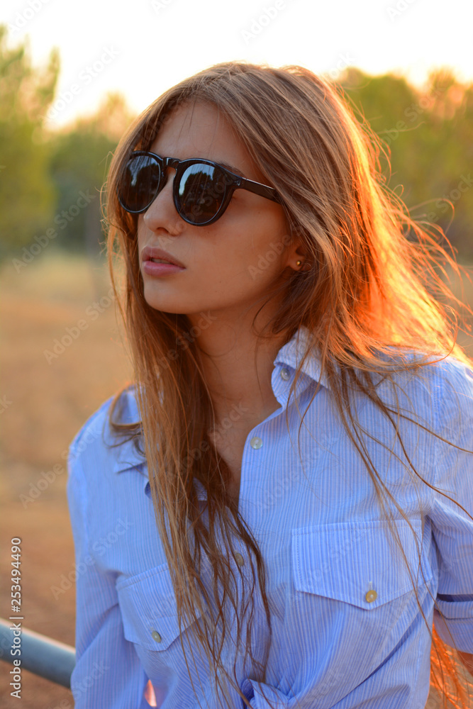 Stylish young hipster woman in black sunglasses in trendy blue shirt posing outdoors a park on a sunny summer day Stylish young hipster woman in black sunglasses in trendy blue shirt posing outdoors a park on a sunny summer day