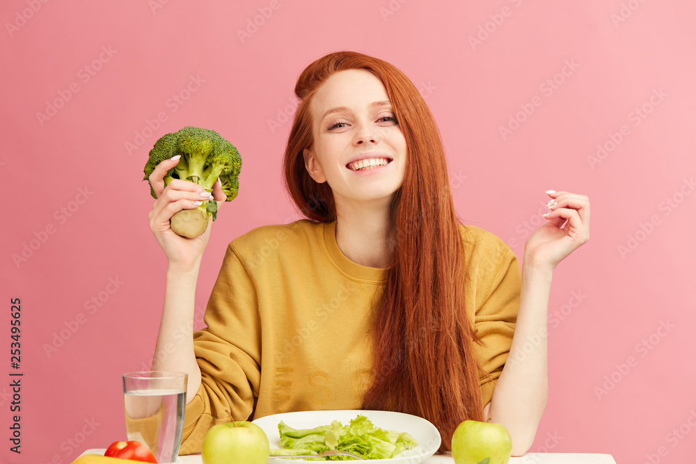 Young beautiful happy attractive female with gorgeous long ginger hair  having dinner with fresh broccoli and vegetables at table, isolated over  pink background. Healthy diet concept. Vegetarian food Stock Photo | Adobe
