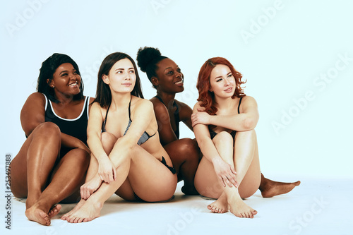 Multiethnic diverse women with cheerful expression smiling and hugging together, looking aside, dressed in underwear, isolated over white background. Interracial lesbian couple.