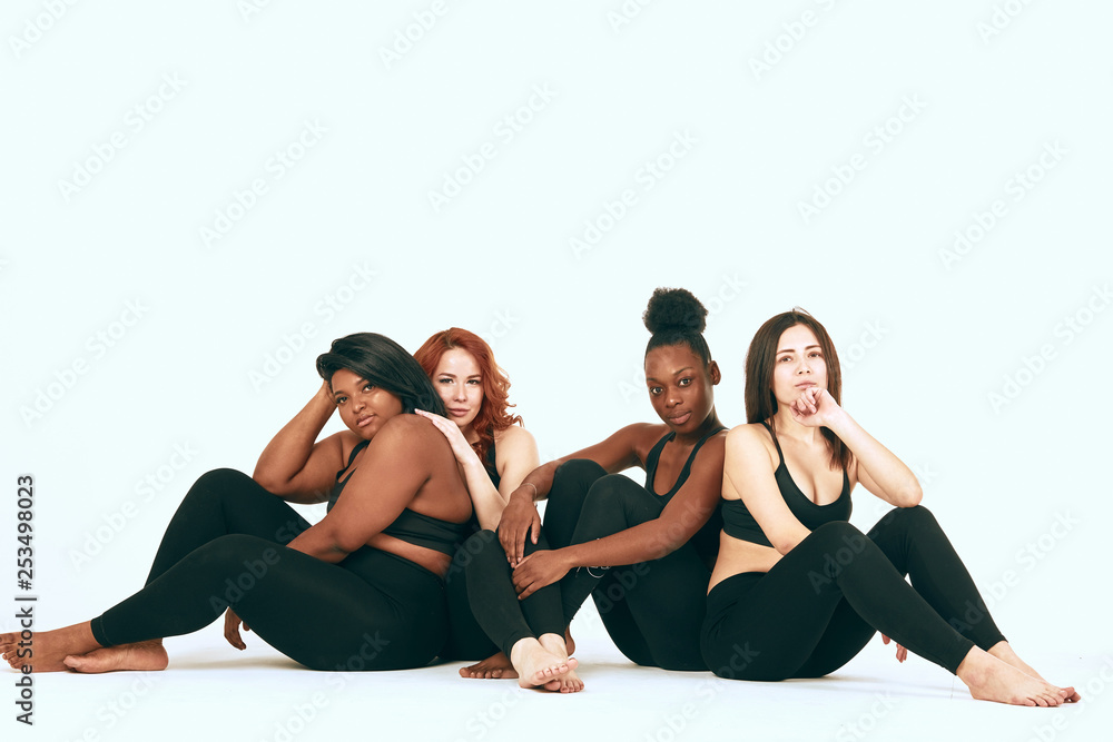 Group of four women of different race, figure type and size in sports outfit sitting back to back together over white background.