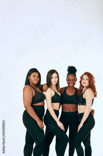 Studio portrait of two dark- skinned and two caucasian women of different figure and size posing together in black sports suits against a gray background.