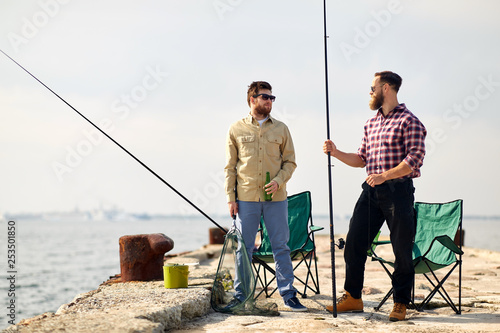 leisure and people concept - happy friends with fishing rods and beer talking on pier at sea