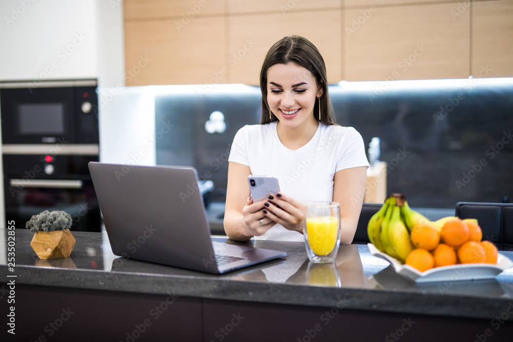Pretty young woman typing on the phone using laptop at home in the kitchen Pretty young woman typing on the phone using laptop at home in the kitchen