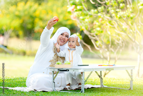 Muslim mother showing a fresh fruit for his little son in the garden on grass field near beautiful lake.