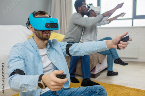Group of multucultural friends having fun at home playing 3D game using hi-tech vitrual reality viewer for gaming. Focus on caucasian man in blue virtual reality headset gesturing as he is driving car