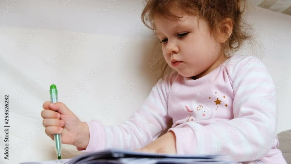 Charming little girl paints a coloring book while sitting on a sofa in her room. The concept of children's educational activities Charming little girl paints a coloring book while sitting on a sofa in her room. The concept of children's educational activities
