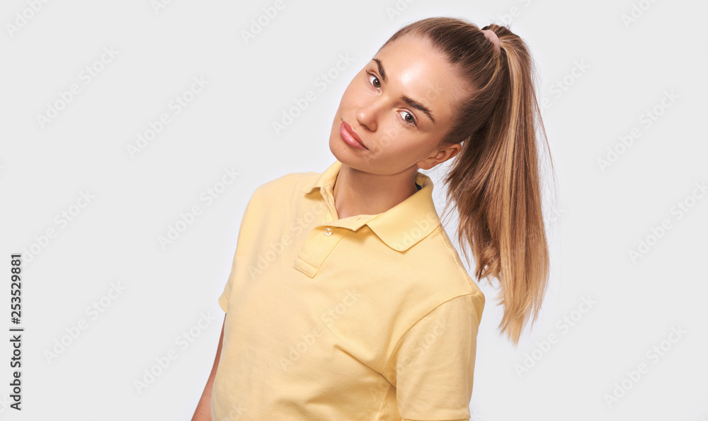 Beautiful blonde young woman with a pony tail looking at camera with cute shy smile, feeling a bit shy or uncomfortable. Pretty female wearing yellow t-shirt on white studio wall. People emotions Beautiful blonde young woman with a pony tail looking at camera with cute shy smile, feeling a bit shy or uncomfortable. Pretty female wearing yellow t-shirt on white studio wall. People emotions