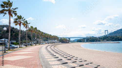 A toll road rest stop in Hakata Island with a row of palm trees by the beachside and a view of the Hakata Oshima Bridge which connects Hakata Island with Oshima Island in the Shikoku Region of Japan.
