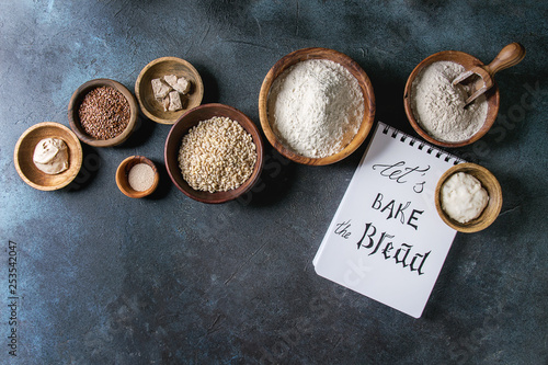 Ingredients for baking bread. Variety of wheat and rye flour, grains, yeast, sourdough and notebook with handwritten lettering over dark blue texture background. Flat lay, space