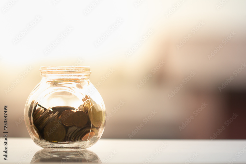 Glass jar / piggy bank full of coins under sunlight on blurred background with copy space. Business, financial, investment, and saving concept Glass jar / piggy bank full of coins under sunlight on blurred background with copy space. Business, financial, investment, and saving concept