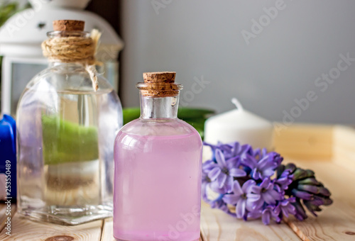 Floral cosmetic water for the face in a transparent bottle stands in the center of the wooden table, in the background there is a transparent white bottle, a purple hyacinth flower