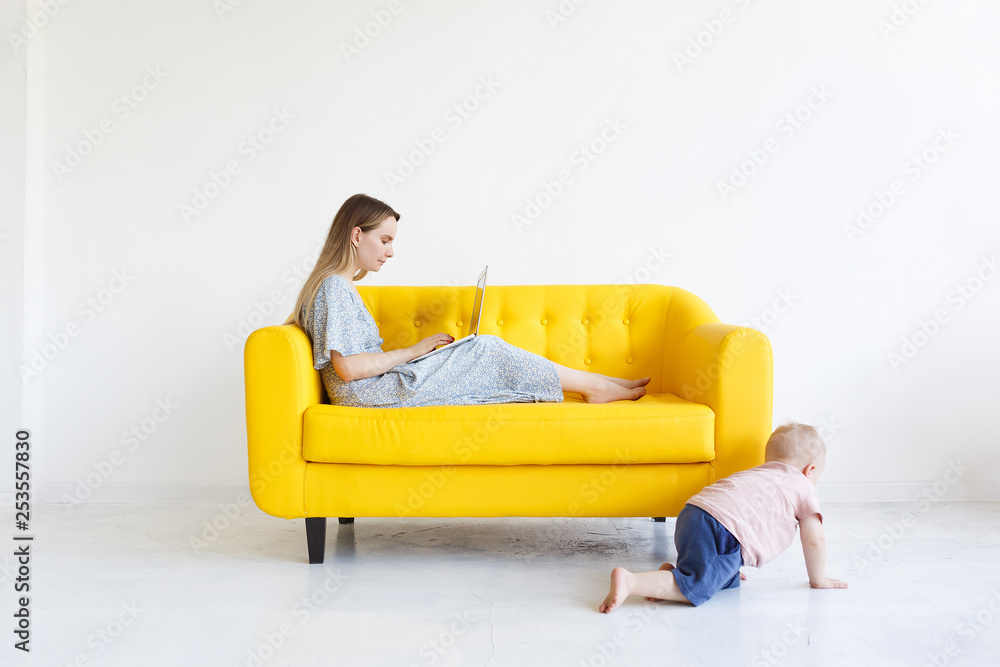 Adorable female freelancer mom in casual dress works remotely on portable laptop computer on yellow sofa, studying forum for mothers, starting business at home while kid is playing alone. Copy space