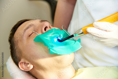 A young man with closed eyes in the dental chair. The dentist works with a dental polymerization lamp.