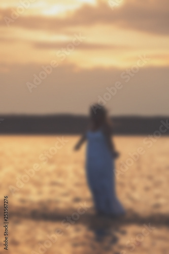 Blurred - blonde girl in a white dress walking on the water at sunset.