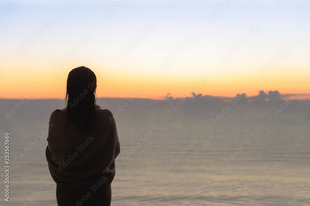 Silhouette of woman with wet hair wrapped in a blanket after swimming. Female in the headphones standing on the beach opposite sea.