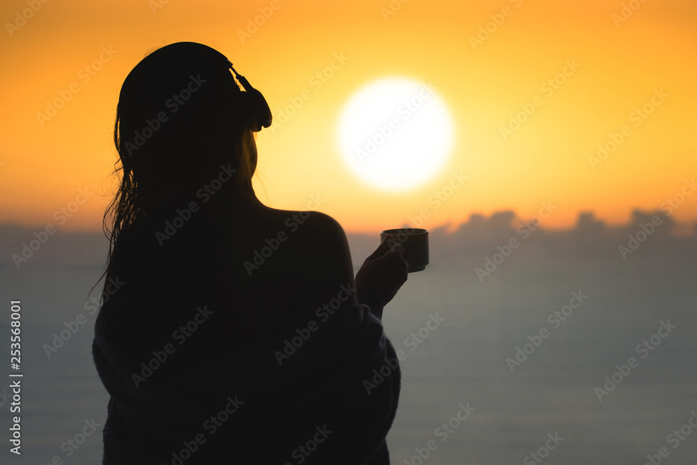 Naked beautiful female with wet hair wrapped in a blanket with cup of coffee standing on the glass balcony early morning at sunrise. Sea view