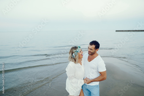 elegant and stylish woman with light curly hair dressed in a white blouse walking on the beach near water along with her elegant man in a white t-shirt and in a blue shorts