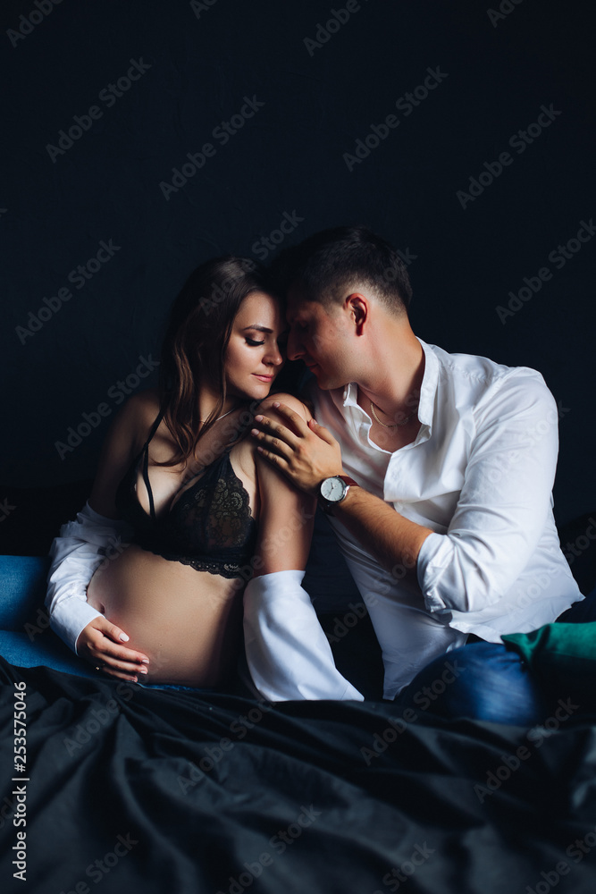 Studio portrait of beautiful and tender couple on bed. Attractive young  pregnant woman in black laced