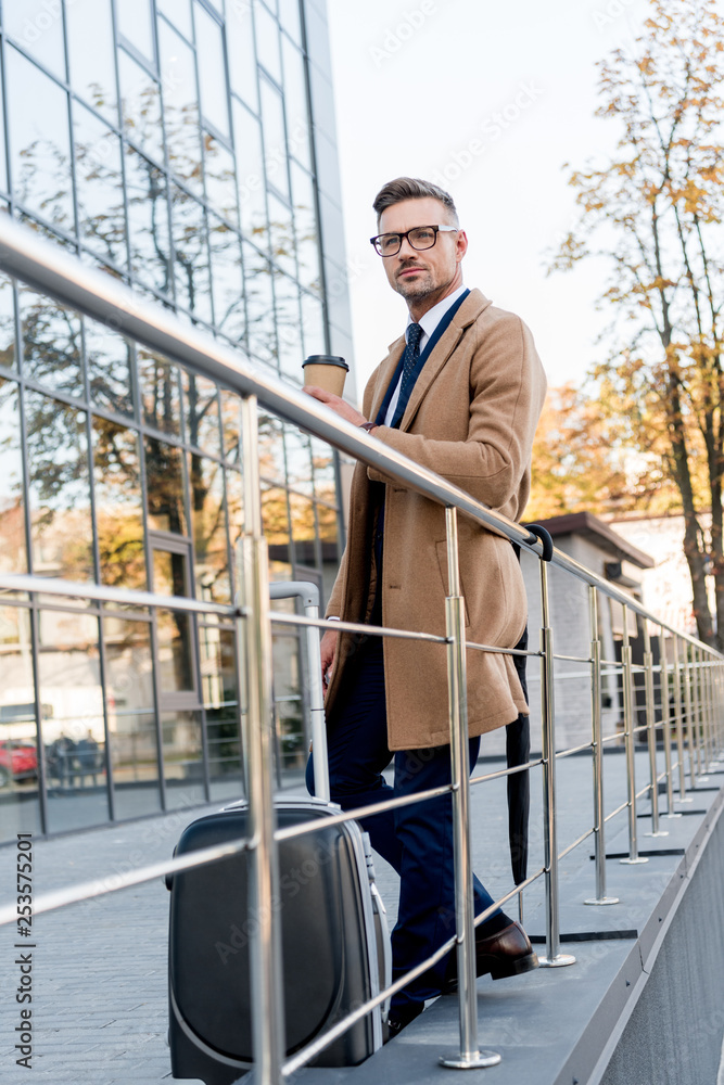 handsome businessman in beige coat holding paper cup while standing near suitcase handsome businessman in beige coat holding paper cup while standing near suitcase