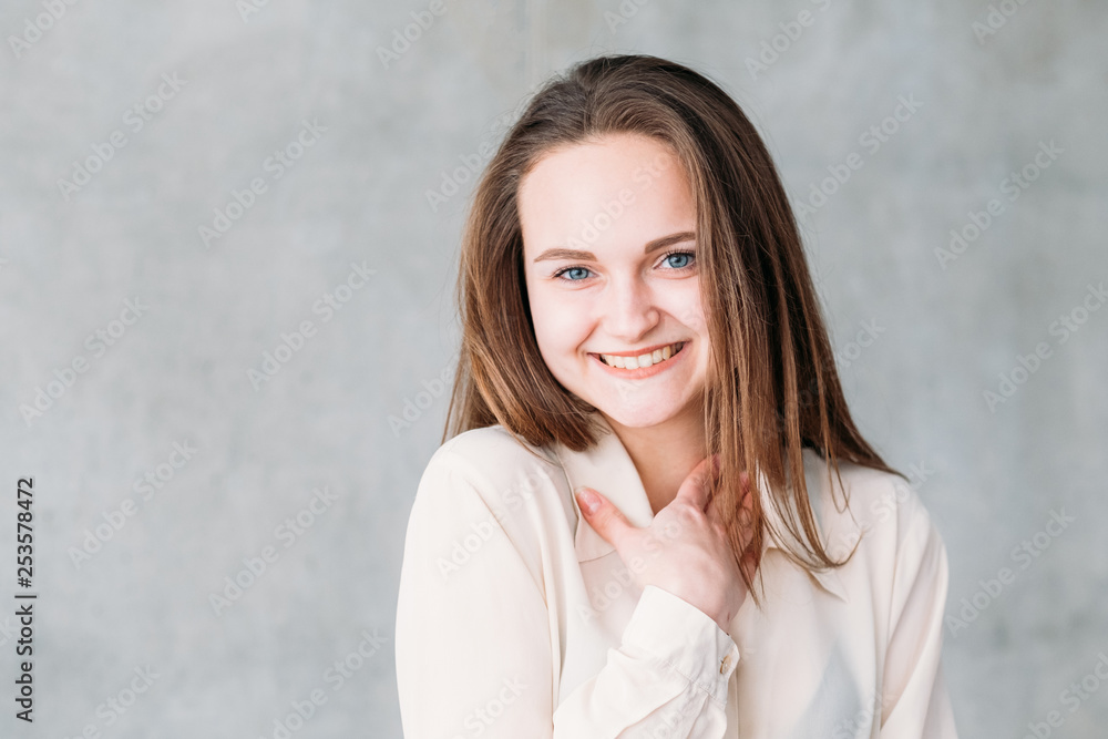 Young woman portrait. Smiling and shy at compliment with hand at neck. Copy space on grey background. Young woman portrait. Smiling and shy at compliment with hand at neck. Copy space on grey background.