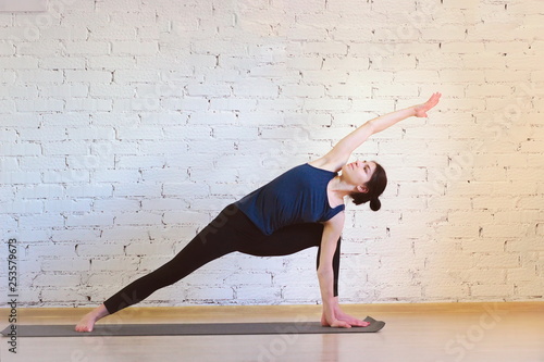 The girl is a professional instructor of hatha yoga practicing asanas in the room against the background of a white brick wall. Utthita Parshvakonasana (extended side angle pose).