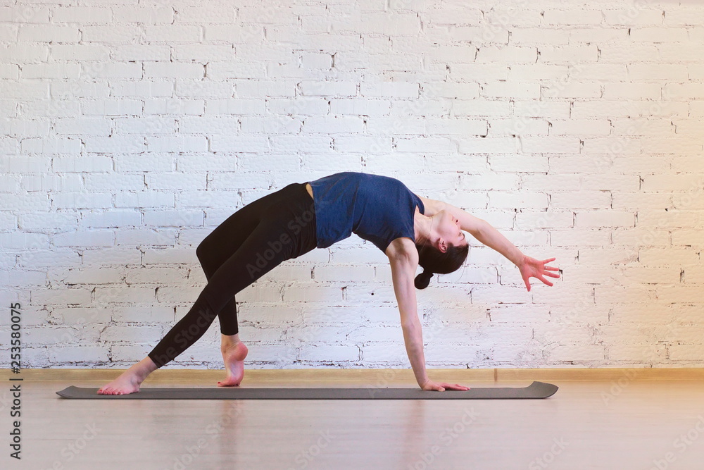 The girl is a professional instructor of hatha yoga practicing asanas in the room against the background of a white brick wall. Camatkarasana (Dancing dog pose or Wild thing).