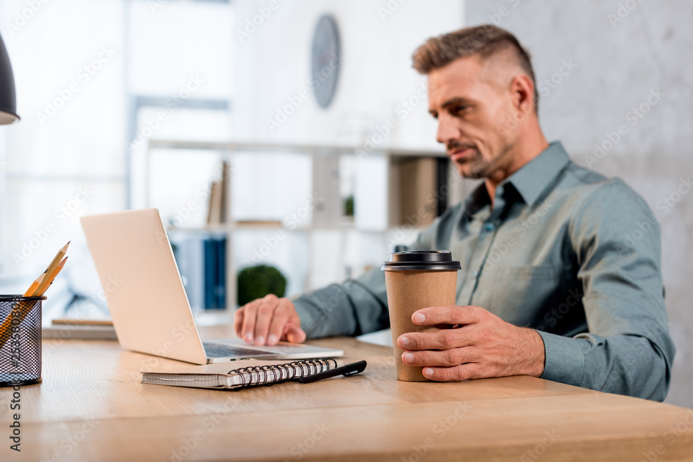 selective focus of paper cup in hand of businessman using laptop in office selective focus of paper cup in hand of businessman using laptop in office