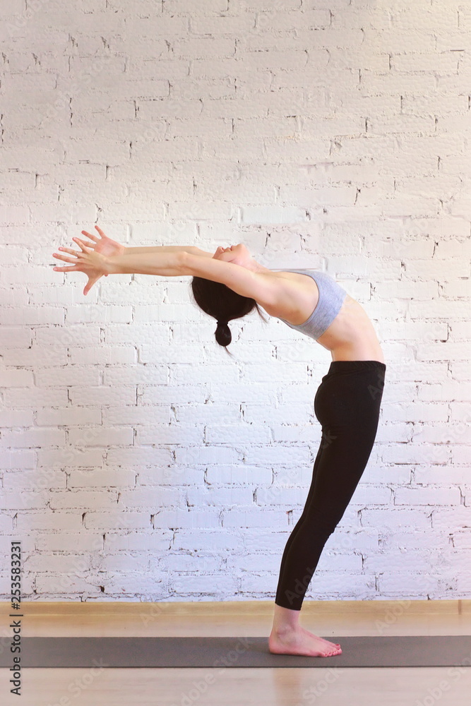 The girl is a professional instructor of hatha yoga practicing asanas in  the room against the background of a white brick wall. Anuvittasana  variation (Standing Backbend). Stock Photo