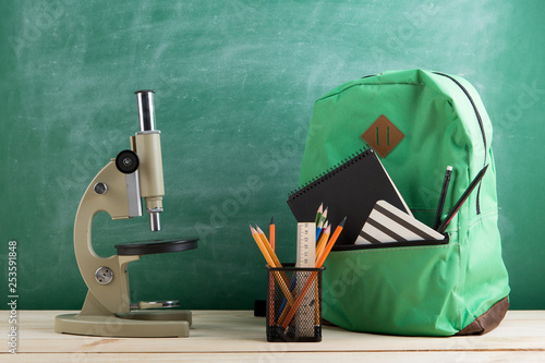 green backpack, microscope, black notebooks and pencils on the background of the blackboard