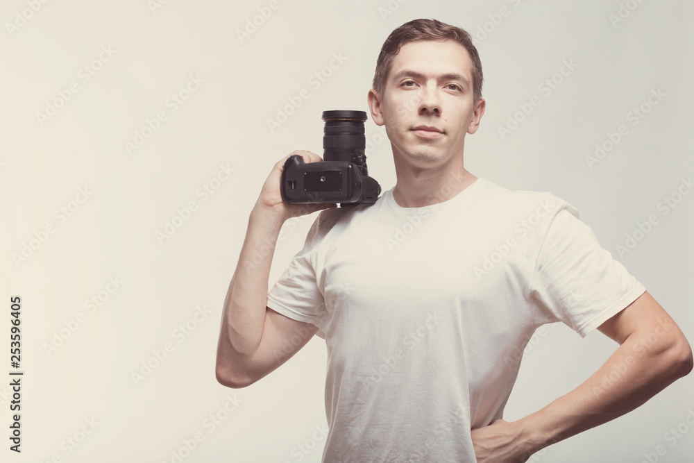 Serious Man with camera isolated on light background. Young man holding digital camera and look on you. Lifestyle, travel and technology concept. Happy boy in white t-shirt love work and dslr camera Serious Man with camera isolated on light background. Young man holding digital camera and look on you. Lifestyle, travel and technology concept. Happy boy in white t-shirt love work and dslr camera