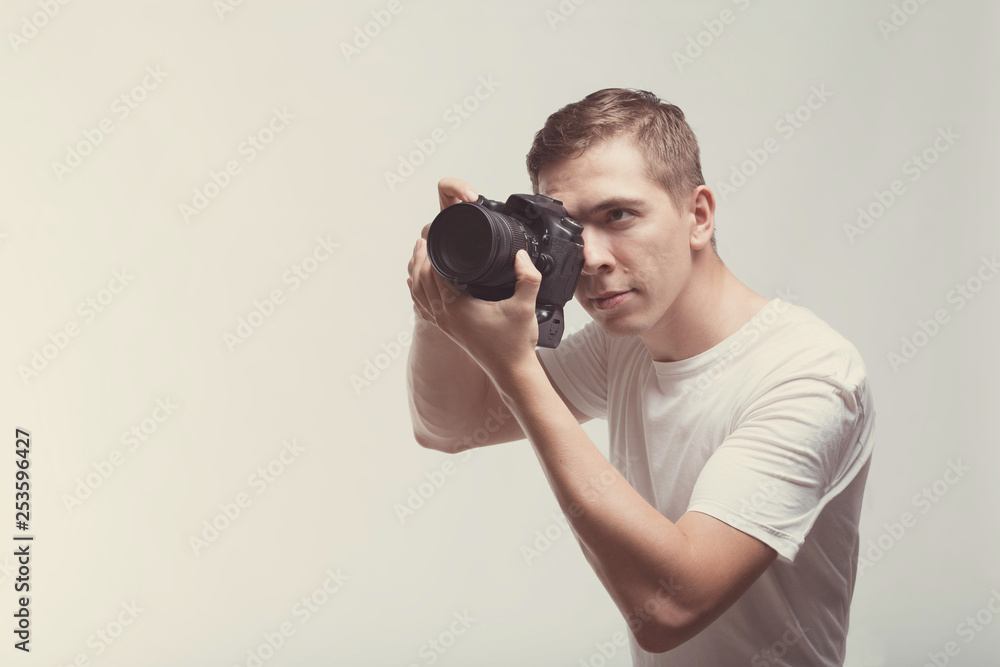 Smiling Man with camera isolated on light background. Young man holding digital camera and making photo looking on side. Lifestyle, travel and technology concept. Happy boy in white t-shirt with dslr