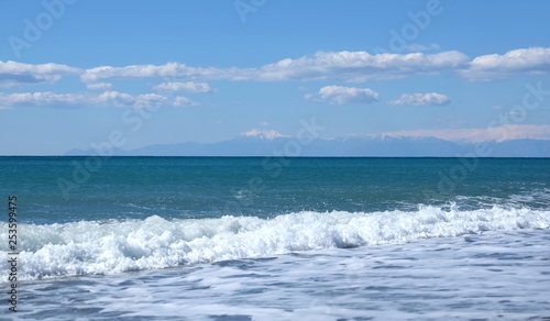 Beautiful landscape with sea, surf with waves with foam runs to long coast and high mountains with snow caps on skyline in haze on sunny summer day