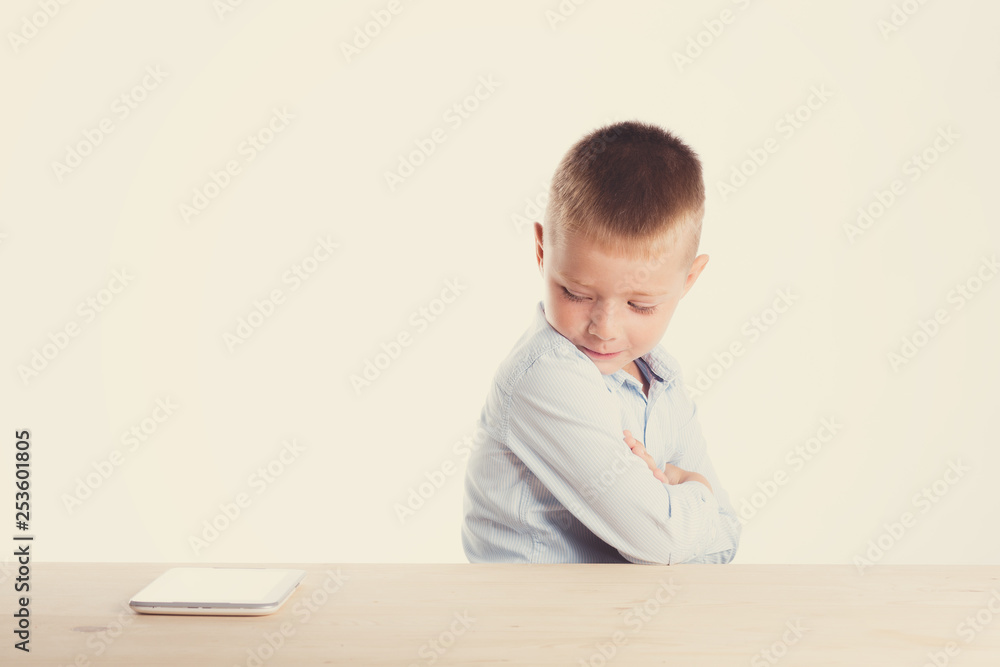 Sad little boy in suit sitting at table with tablet gadget. Children portrait. Back to school. Stylish man in fashionable suit shirt and pants with blue eyes Sad little boy in suit sitting at table with tablet gadget. Children portrait. Back to school. Stylish man in fashionable suit shirt and pants with blue eyes