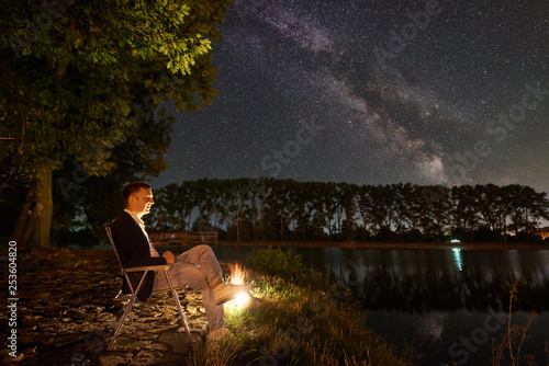 Man tourist having a rest on lake shore near campfire, sitting on chair under big tree, enjoying beautiful view of night sky full of stars and Milky way, city lights on background.