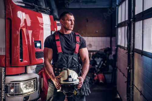 Fototapeta Muscular fireman holding a protective helmet in a garage of a fire department, s
