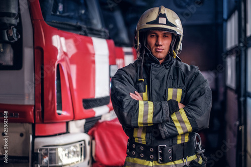 Confident fireman wearing protective uniform standing next to a fire engine in a garage of a fire department, crossed arms and looking at a camera photo