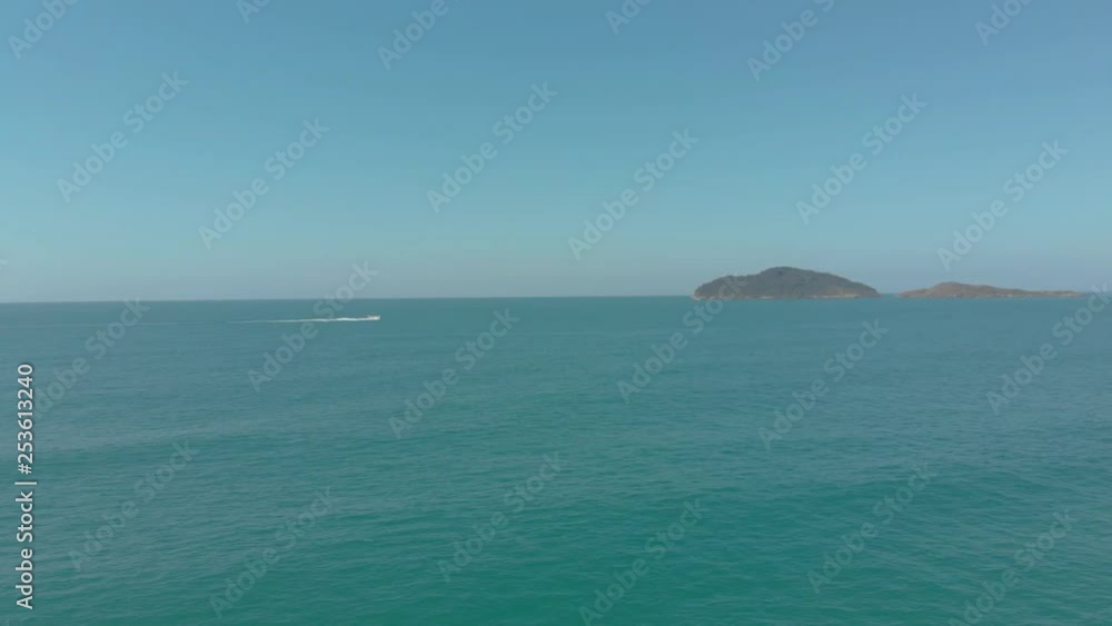 Drone shot of the calm, clear and crystal ocean near Santinho Beach, Florianópolis, Brazil, with a little hilly island full of trees, a boat passing by, on a sunny day Drone shot of the calm, clear and crystal ocean near Santinho Beach, Florianópolis, Brazil, with a little hilly island full of trees, a boat passing by, on a sunny day
