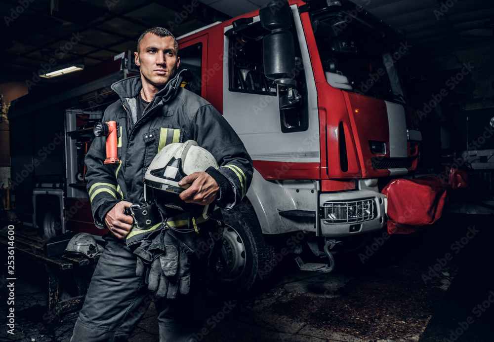 Fireman wearing uniform holding a helmet and looking at a camera while standing near a fire truck in a garage of a fire department at night. Fireman wearing uniform holding a helmet and looking at a camera while standing near a fire truck in a garage of a fire department at night.