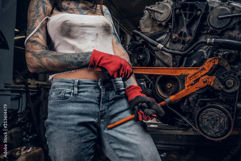 Cropped image. Stylish female model with tattoed body repairs a car engine suspended on a hydraulic hoist in the workshop.