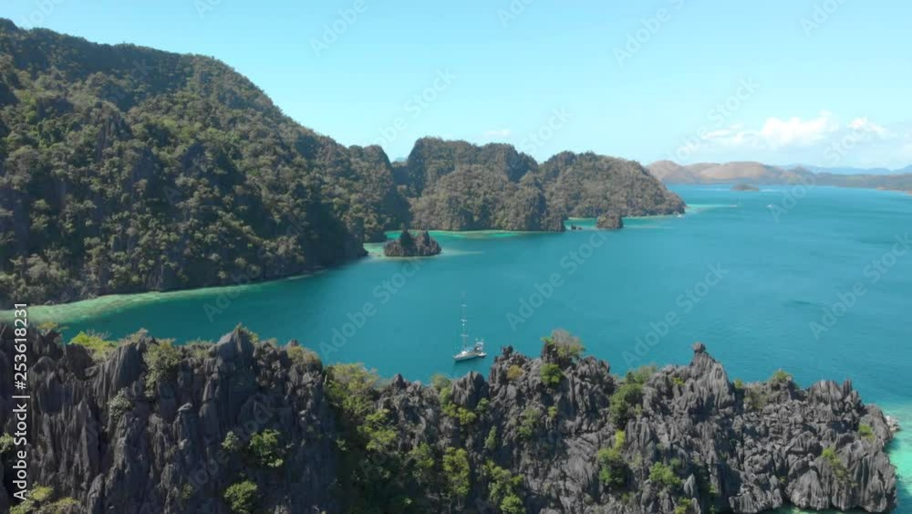 Steep rugged islands of Busuanga, Philippines covered in lush green trees and surrounded by clear turquoise waters. Aerial view from drone ascending revealing a tall ship anchored in a peaceful bay. Steep rugged islands of Busuanga, Philippines covered in lush green trees and surrounded by clear turquoise waters. Aerial view from drone ascending revealing a tall ship anchored in a peaceful bay.