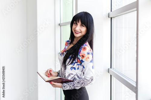 Young Beautiful Businesswoman standing near the window, writing in a notebook, looking at the camera smiling.