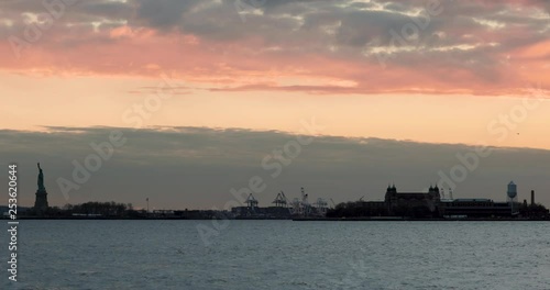 Static shot of the Statue of Liberty and Ellis Island silhouetted against sunset skies.