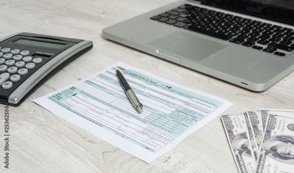 US tax form next to computer laptop, dollar bills, calculator and tax form 1040. tax form us business income office hand fill concept US tax form next to computer laptop, dollar bills, calculator and tax form 1040. tax form us business income office hand fill concept