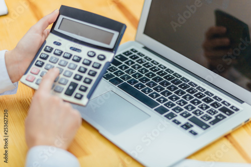 Close up of business woman holding hands with black calculator and laptop while sitting at desk in office background.