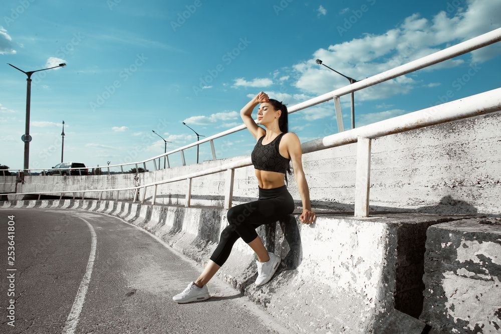 Portrait of beautiful brunette. Sexy girl rest after running. Female fitness model working out outdoor in city. Young woman jogger athlete training on road. Concept of healthy lifestyle.
