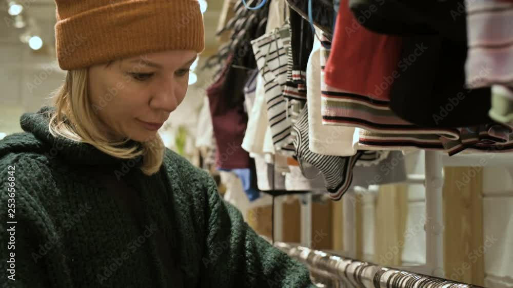 A girl in a green sweater and a yellow hat walks through a store of things and chooses what to buy. Touches things on hangers and looks at price tags A girl in a green sweater and a yellow hat walks through a store of things and chooses what to buy. Touches things on hangers and looks at price tags