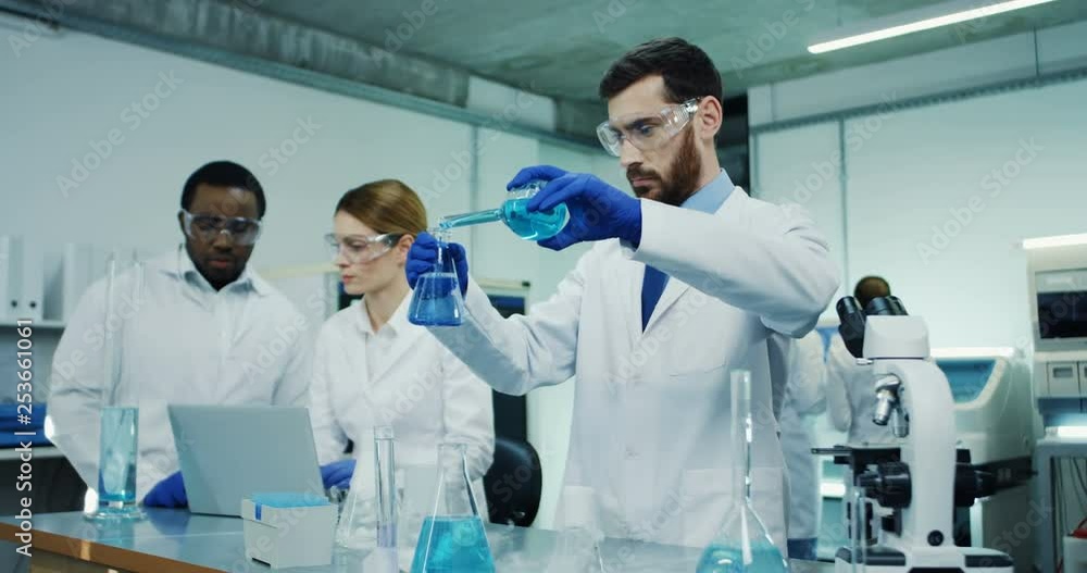 Caucasian male scientist in the glasses and white robe doing some research with the test tubes in the laboratory room. Mixed-races male and female colleagues on the background. Caucasian male scientist in the glasses and white robe doing some research with the test tubes in the laboratory room. Mixed-races male and female colleagues on the background.
