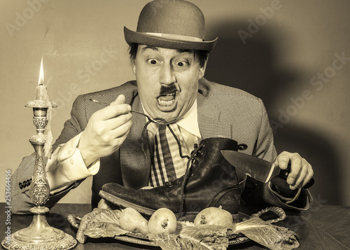 Poverty stricken, bankrupt, poor Caucasian male with mustache ,eating a boot by candle light , in a dirty room ,dressed as Charlie Chaplin type character ,wearing a hat.