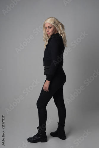  full length portrait of a blonde girl wearing modern black jacket and pants, standing pose facing away from the camera on grey studio background.