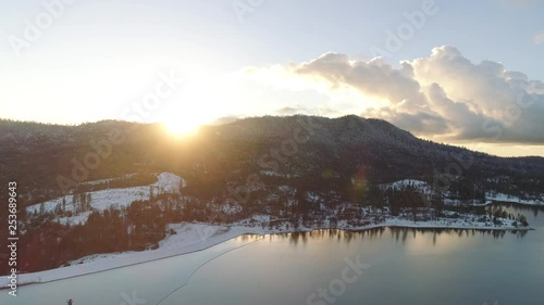 Dazzling aerial shot turning over a lake in winter with bright sun and lens flare. Snowy ground and hills.